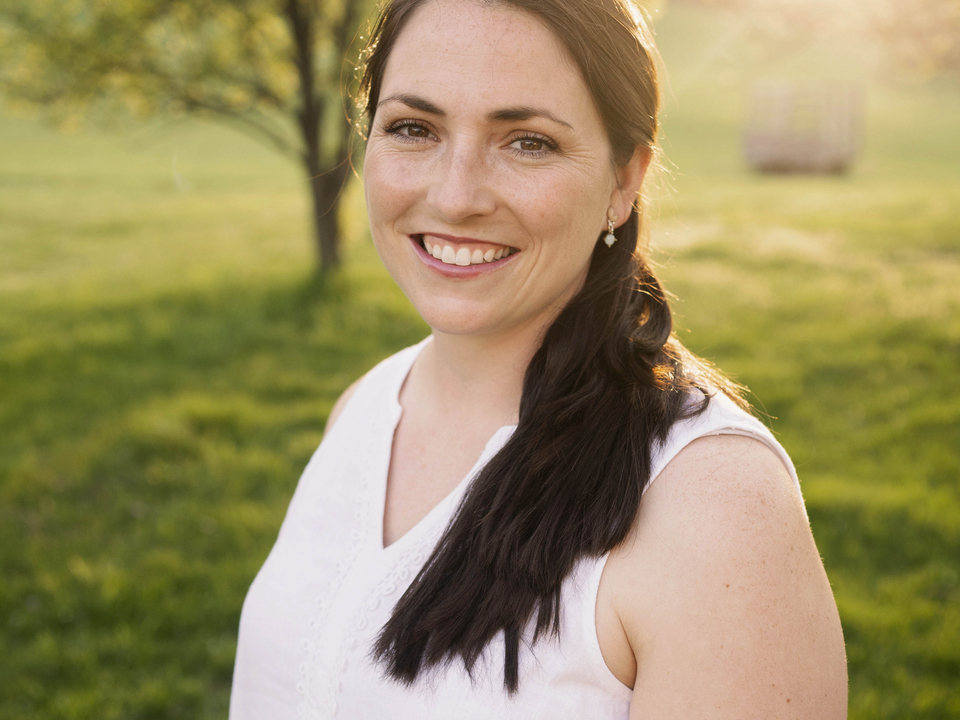 Marie Miller dressed in white sleeveless top in a field of grass with tree and sun in background. 