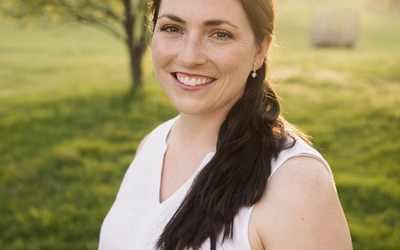 Marie Miller dressed in white sleeveless top in a field of grass with tree and sun in background. 