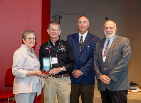 Dr. Brenda McCartney, Chuck Trautwein, Dr. David Nelson, and Dr. Bill Dennison standing together, facing camera, with dark red wall behind them. Chuck is holding the glass plaque award. 