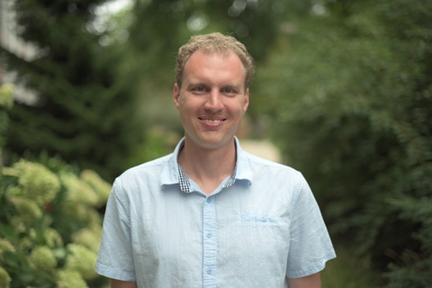 Headshot of Adam Benfield in light blue, short sleeved button-down shirt with blurred green foliage in background.  